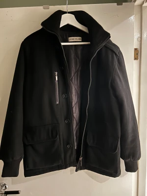 Svart jacka från Stone Island - Stilren svart jacka från Stone Island med hög krage, dragkedja och knappar framtill. Jackan har två stora fickor med lock och en bröstficka med dragkedja. Quiltat innerfoder och ribbade muddar vid ärmslut. Perfekt för kyligare dagar.
