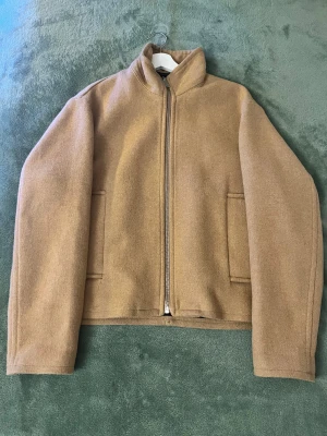 Beige ulljacka med dragkedja - Snygg beige jacka i ull med rak passform och hög krage. Jackan har två diskreta fickor framtill och stängs med en dragkedja. Perfekt för dig som gillar stilrena och minimalistiska plagg med en modern touch. Sparsamt använd och mycket bra skick. M men passar S och L Nypris 800kr 