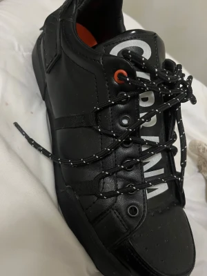 Svarta sneakers från Dolce & Gabbana - Snygga svarta sneakers från Dolce & Gabbana med vit logga på plösen och orange innersula. Skorna har svarta snören med vita detaljer, perforerad tå och platt sula med D&G-mönster undertill. Tillverkade i skinn för en lyxig känsla.