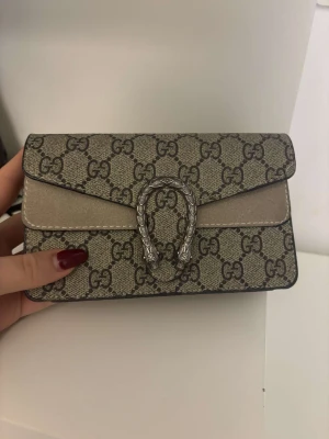 MINI Beige kuvertväska med kedja - Snygg kuvertväska i beige med klassiskt GG-mönster och silverfärgad kedja. Väskan har en unik metallspänne framtill och detaljer i mocka. Perfekt storlek för det viktigaste och ger en lyxig vibe till din outfit.