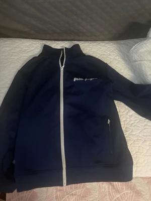 Blå track jacket från Palm Angels - Snygg marinblå track jacket från Palm Angels med vit dragkedja och logga på bröstet. Jackan har hög krage, långa ärmar och fickor med dragkedja. Perfekt för en sportig och trendig look.