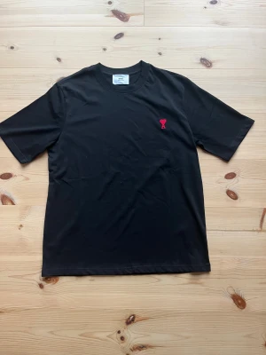Svart t-shirt från Ami med rött hjärta - Svart t-shirt från Ami Alexandre Mattiussi med klassisk passform och rund hals. På bröstet finns ett broderat rött hjärta med bokstaven A. Materialet är mjuk bomull och t-shirten har korta ärmar. Perfekt för dig som gillar stilrena detaljer.