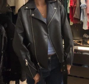 Svart skinnjacka med dragkedjor - Säljer en svart skinnjacka med coola silverfärgade dragkedjor och detaljer. Jackan har klassisk bikerstil med bred krage, fickor med dragkedja och bälte nedtill. Passformen är loose och den har långa ärmar med dragkedjor vid ärmslut. Perfekt för dig som vill ha en edgy look.