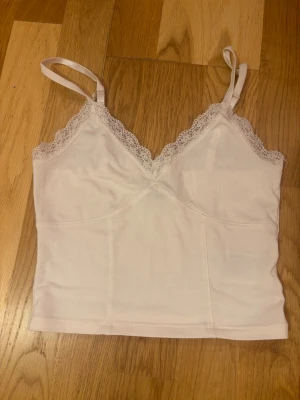 Vit spetskantad linnetopp - Säljer en ljusvit linnetopp med smala axelband och v-ringad hals. Toppen har fina spetsdetaljer längs halsringningen och är croppad i modellen. Materialet är mjukt och känns skönt mot huden, perfekt för varma dagar eller att styla lager på lager.