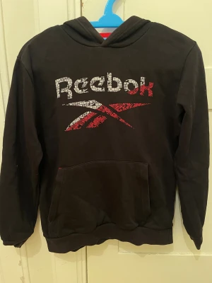 Svart hoodie från Reebok - Svart hoodie från Reebok. Tröjan har huva med röd insida och Reebok-tryck på snörena. Passar 11-12Y.