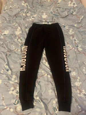 Svarta Hoodrich mjukisbyxor med tryck - Svarta mjukisbyxor från Hoodrich med vit logotyptryck längs båda benen. Byxorna har smal passform, ribbade muddar vid benslut och blå piping längs sidorna. Utrustade med snörning i midjan för extra komfort. Perfekta för chill eller streetwear.