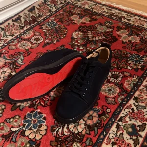 Christian Louboutin svarta sneakers - Snygga svarta sneakers från Christian Louboutin i mocka med ikonisk röd sula. Skorna har rund tå, svarta snören och diskret broderad logga på hälen. Insidan är i beige läder för extra lyxig känsla. Perfekta för dig som vill sticka ut med stil.