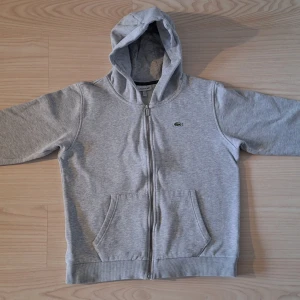 Grå hoodie från Lacoste med dragkedja - Passar för 12 år, inte 14 som det står i etiketten, tyvärr lite krymt i tvätten Annars i gott skick, inga tecken på slitage. Nypris på kidsbrandstore 1199 kr. Snygg grå hoodie från Lacoste med klassisk krokodillogga på bröstet. Tröjan har huva, hel dragkedja och två stora fickor framtill. Perfekt för chill dagar och enkel att matcha med det mesta. Mjuk och skön bomullsblandning.