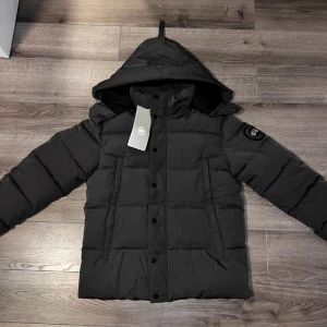 Svart pufferjacka från Canada Goose helt ny - Säljer en svart pufferjacka från Canada Goose med huva och klassisk patch på ärmen. Jackan har långa ärmar, knappar och dragkedja framtill samt en clean, boxig passform. Perfekt för kalla dagar och riktigt snygg till streetwear-stilen.