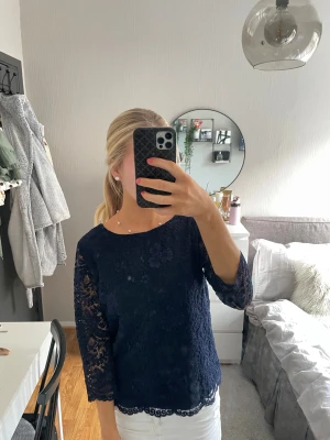 Mörkblå spetsblus med trekvartsärm - Säljer en mörkblå blus i spets med blommigt mönster och fina detaljer längs nederkanten. Blusen har trekvartsärmar och är lätt transparent på ärmarna. Perfekt för dig som gillar en feminin och stilren look.