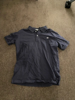 Mörkblå pikétröja från GANT 150 - Snygg mörkblå pikétröja från GANT med klassisk krage och två knappar framtill. Tröjan har korta ärmar och ett broderat GANT-emblem på bröstet. Tillverkad i mjuk bomull som är skön mot huden. Perfekt för dig som gillar stilren och enkel design.