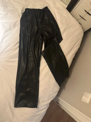 Svarta skinnbyxor från NA-KD - Säljer ett par svarta skinnbyxor från NA-KD i storlek 34. Byxorna har elastisk midja och raka ben med loose passform. Materialet är syntetiskt skinn som ger en snygg och edgy look. Perfekta för dig som vill sticka ut med en cool vibe.
