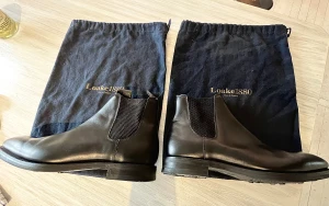 Svarta Chelsea boots från Loake 1880 - Säljer ett par oanvända klassiska svarta Chelsea boots från Loake 1880, modell Chatsworth med etikett kvar, Skorna är i slätt skinn med elastiska paneler på sidorna och robust Dainite gummisula för extra grepp. Medföljer original dustbags. Perfekta för dig som vill ha stilrena boots med brittisk känsla.