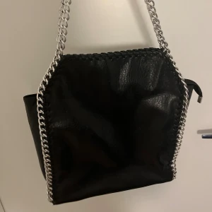 Svart axelväska med kedjedetaljer - Cool svart axelväska i skinnimitation med grova silverfärgade kedjor längs kanterna och som axelrem. Väskan har en fyrkantig form och dragkedja baktill för extra förvaring. Perfekt för dig som gillar edgy och stilren design.