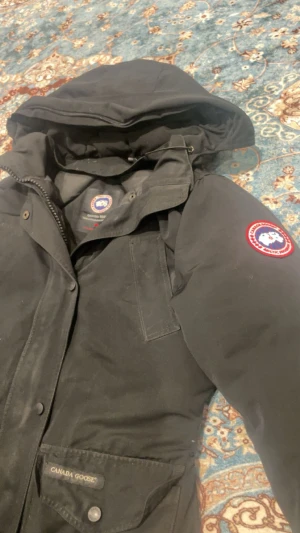 Svart Canada Goose parkajacka M passar S och Xs äkta  - Svart parkajacka från Canada Goose med klassisk logga på ärmen och stora fickor framtill. Jackan har huva, dragkedja och knappar, samt en stilren design som är perfekt för kalla dagar. Materialet är slitstark polyester och jackan är långärmad.
