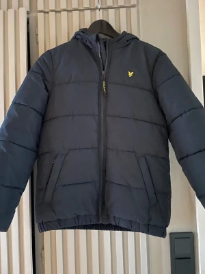 Mörkblå pufferjacka från Lyle & Scott - Snygg mörkblå pufferjacka från Lyle & Scott med  luva. Den är använd ungäfer 2-3 gånger så nästan helt oanvänd, den passar 13 år men den skulle sitta bra med 14 år också         