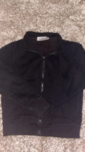  zipjacka från Stone Island - Svart zipjacka från Stone Island i storlek M. Jackan har en stilren design med ståkrage, hel dragkedja och två fickor framtill. Tillverkad i mjukt bomullsmaterial som är skönt att bära. Perfekt för dig som gillar minimalistisk och clean streetwear.