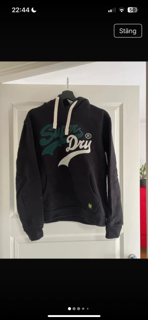Svart Superdry hoodie med tryck - Svart hoodie från Superdry i storlek M med stort broderat logotryck i grönt och vitt på bröstet. Tröjan har huva med vita snören och en klassisk magficka. Materialet är mjuk bomull och polyester, perfekt för chill dagar.