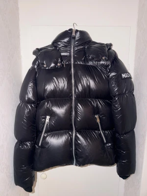 Svart pufferjacka från Mackage - Snygg svart pufferjacka från Mackage med glansig finish, hög krage och avtagbar huva. Jackan har två dragkedjeförsedda fickor framtill och tydlig logga på ärmen. Perfekt för kalla dagar när du vill hålla stilen.  Storlek 38 vilket motsvarar M/L