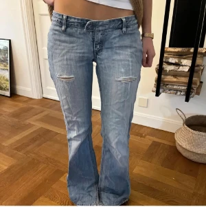 Ljusblå bootcut jeans med slitningar  - Säljer ett par ljusblå jeans med bootcut-modell och låg midja. Jeansen har slitna detaljer vid låren. Och även på bakre fickan. Ena knappen är borta framtill men enkelt att sätta på en egen eller bara använda skärp till dom🥰🥰
