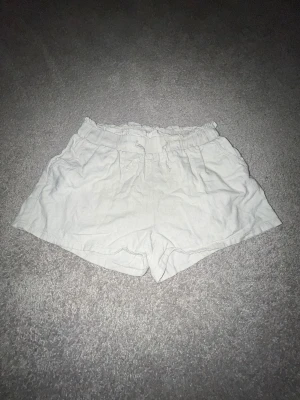 Beigea linneshorts  - Fräscha beiga barnshorts från Lindex i storlek 146. De har en högmidjad passform med elastisk midja och snörning framtill. Materialet är mjukt och luftigt linne, perfekt för varma dagar. Shortsen har fickor på sidorna och en enkel, clean look.