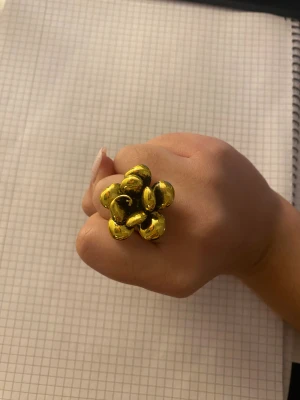 Guldig blomformad ring - Unik ring i guldton med stor blomformad design. Blomman har flera blad med rundade former och en glansig finish som verkligen sticker ut. Perfekt för dig som vill ha ett statement piece med cool vibe och mycket attityd.