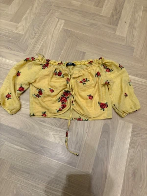 Gul blommig croppad blus från H&O - Superfin gul croppad blus från H&O med broderade röda rosor och gröna blad. Blusen har offshoulder-modell, långa puffärmar och snörning framtill. Perfekt för dig som vill sticka ut med en färgglad och romantisk vibe.