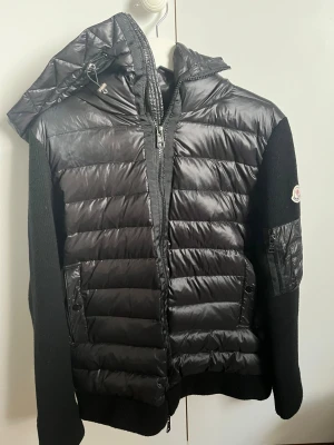 Svart Moncler pufferjacka med huva - Snygg svart pufferjacka från Moncler med stickade ärmar och quiltad framsida. Jackan har dragkedja, huva och en ficka med dragkedja på ärmen. Perfekt för kyliga dagar. Knappt använd och säljer pågrund av storlek och passform. Skavank i fickan 