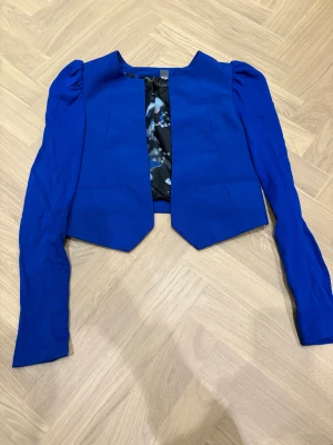 Kornblå kavaj med puffärm - Snygg kornblå kavaj med lång puffärm och coolt mönstrat foder i blått, svart och gult. Jackan har en öppen front och markerade axlar för en trendig look. Passar dig som vill sticka ut med färg och stil.