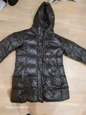 Svart dunjacka från Uniqlo med huva - Säljer en svart dunjacka från Uniqlo i storlek M. Jackan har en snygg quiltad design, huva och dragkedja framtill. Den är lång och har två praktiska fickor på sidorna. Perfekt för kalla dagar och har en stilren look som funkar till det mesta.