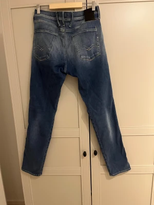 Blå Replay Anbass jeans tapered fit - Säljer ett par blå Replay Anbass jeans med snyggt slitna detaljer och klassisk femficksdesign. Jeansen har tapered fit, normal passning och är tillverkade i mjukt jeansmaterial. Perfekt för dig som gillar en modern och avslappnad stil.