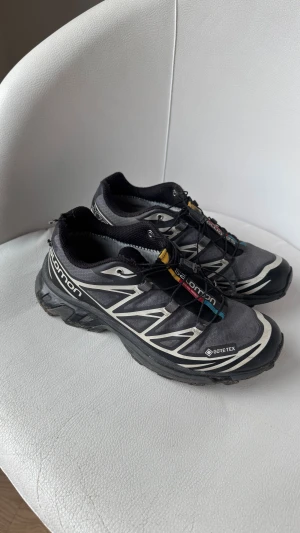 Salomon XT-6 Gore-Tex sneakers svart - Salomon XT-6 sneakers i svart med vita och grå detaljer, snabbsnörning och Gore-Tex-material. Skorna har en sportig design med robust sula och färgdetaljer i gult, rött och blått på plösen. Perfekta för dig som gillar outdoor och streetstyle.