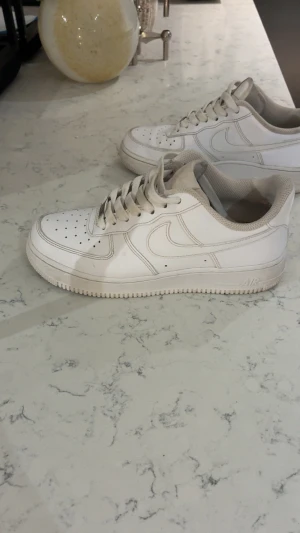 Nike Air Force 1 vita sneakers - Klassiska Nike Air Force 1 sneakers i helvitt skinn med perforerad tå och rund sula. Lågt skaft, snörning och ikonisk Swoosh-logga på sidan. Skorna har en platt sula och meshfoder runt ankeln för extra komfort. Perfekt för dig som gillar streetstyle. Har en liten skada där inne i hälen som visas i bilderna! Men det är inget som syns när man har på sig det 