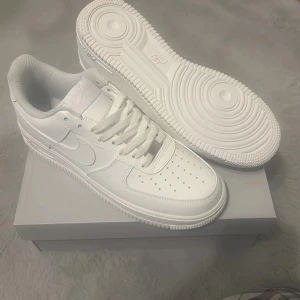 Nike Air Force 1 helvita sneakers - Helt nya och oanvända Nike Air Force 1 sneakers i helvit färg med klassisk låg siluett. Skorna har rund tå, snörning och perforerad tåbox för extra ventilation. Tillverkade i skinn med vit yttersula och ikonisk Nike Swoosh på sidan. Perfekt för dig som gillar clean och tidlös streetstil.