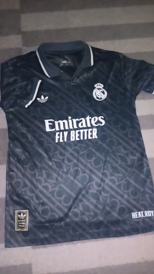 (Äkta 25/26) Real Madrid Bellingham #5 Adidas tröja - Snygg svart Real Madrid fotbollströja från Adidas med Bellingham och nummer 5 på ryggen. Tröjan har korta ärmar, krage med vit kant och ett diskret mönster över hela tyget. Materialet är lätt och andas, perfekt för match eller träning.