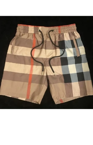 Rutig Burberry shorts med snörning - Snygga shorts från Burberry med klassiskt rutigt mönster i beige, svart, vit och orange. De har elastisk midja med svart snörning och är tillverkade i ett lätt och bekvämt material, perfekt för varma dagar. Ikonisk design som sticker ut.