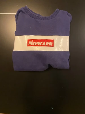Blå Moncler sweatshirt med logga - Säljer en blå sweatshirt från Moncler med en bred vit rand över bröstet och röd broderad Moncler-logga. Tröjan har rund halsringning och är tillverkad i mjuk bomull. Perfekt för dig som gillar streetwear och vill sticka ut med en snygg logga. Storlek 12A, upplevs som liten i storlek.