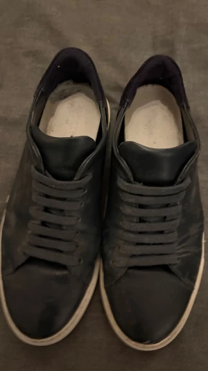 Mörk blåa sneakers från Axel Arigato - Stilrena svarta sneakers från Axel Arigato i skinn med klassisk vit sula. Modellen har rund tå, platt sula och snörning. Perfekta för dig som gillar minimalistisk och clean design. Skorna kommer med originalkartong och dustbag.