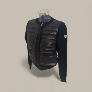 Svart quiltad cardigan jacka Moncler - Svart cardigan jacka från Moncler med quiltad, glansig front och två fickor med knappstängning. Jackan har långärmade stickade ärmar och rund hals. Dragkedja framtill och Moncler-logga på vänster ärm. Perfekt för dig som gillar stilren och sportig look.