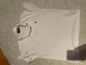 Vit pikétröja Polo Ralph Lauren M - Vit pikétröja från Polo Ralph Lauren i custom slim fit. Klassisk design med korta ärmar, krage och två knappar framtill. Broderad marinblå logga på bröstet. Tillverkad i bomull med en lätt textur som känns fräsch och stilren. Pris är inte hugget i sten, bara lite rynkor med stryker den innan den skickas.