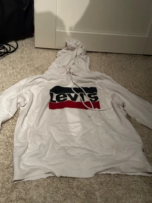 Vit Levi's hoodie med logga, strl M - Säljer en vit hoodie från Levi's i storlek M med stor, klassisk logga i svart, rött och mörkblått på bröstet. Hoodien har snörning i huvan och en stor magficka. långärmad modell, perfekt för chill dagar.
