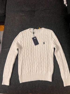 Vit kabelstickad tröja Polo Ralph Lauren - Snygg vit kabelstickad tröja från Polo Ralph Lauren med rund hals och klassisk broderad logga på bröstet. Tröjan har långa ärmar och ribbade muddar vid ärmslut och nederkant. Perfekt för dig som gillar stilrena och tidlösa plagg.