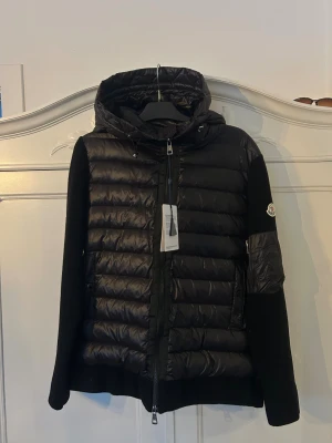 Moncler Cardigan - Svart pufferjacka från Moncler med stickade långärmade ärmar och quiltad kropp. Jackan har huva med snörning, dragkedja framtill och fickor med dragkedja. Moncler-logga på ärmen och extra ficka med dragkedja på ena ärmen. Snygg, modern och perfekt för kyliga dagar. Säljer pågrund av fel storlek. 