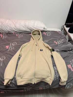 Beige hoodie från Stone Island L - Säljer en beige hoodie från Stone Island i storlek L. Tröjan har huva med snörning, dragkedja framtill och den klassiska Stone Island-loggan på vänster ärm. Mjuk insida och två fickor framtill. Perfekt för chill dagar och streetwear. Den har aldrig blivit änvänd har bara hängt den. Anledningen att jag säljer den är att den är för stor när jag beställde den.