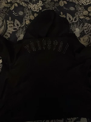 Svart vindjacka från Trapstar - Säljer en svart vindjacka från Trapstar med huva och snyggt tryck på ryggen. Jackan har en glansig finish, dragkedja framtill och en rund logodetalj på sidan. Perfekt för dig som vill ha streetstyle med edge.