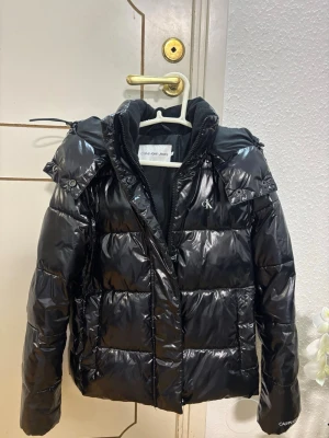 Svart pufferjacka Calvin Klein Jeans XS - Svart glansig pufferjacka från Calvin Klein Jeans i storlek XS. Jackan har huva, dragkedja med CK-detalj och logga på bröstet. Fodrad insida och quiltad design som ger en schysst look. Perfekt för kalla dagar när du vill vara både varm och snygg. Använt den en biter förra året. Jätte snygg på! Passar även de som har storlek S.!😍😍köptes från Zalando:)