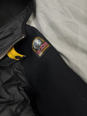 Svart pufferjacka från Parajumpers - Svart pufferjacka från Parajumpers med huva och coolt gult band vid kragen. Jackan har quiltade paneler, dragkedja framtill och en snygg badge på axeln. Materialmix med polyester och syntet, perfekt för dig som gillar streetstyle.