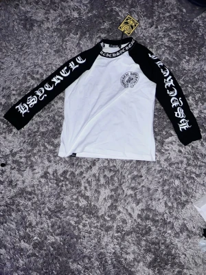 Chrome hearts shirts - Säljer en långärmad t-shirt från Hyraw med vit kropps och svarta ärmar. Tryck i gotisk stil på ärmarna och runt halsen, samt en liten logga på bröstet. Skön passform och cool streetwear-vibe, perfekt för dig som gillar edgy stil.