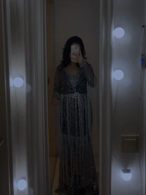 Oanvänd Glittrig långklänning med fransar  - Säljer en maxad långklänning i svart och silver med glittriga detaljer, transparenta ärmar och v-ringning. Klänningen har ett bälte i midjan och långa fransar som ger extra effekt. Perfekt för dig som vill sticka ut med ett unikt mönster och mycket glitter och funkar bra till bröllop och fester 🥳😍❤️❤️‍🔥