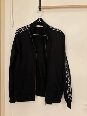 Svart Givenchy hoodie med dragkedja - Svart hoodie från Givenchy med vit logotyp längs axlar och ärmar. Tröjan har dragkedja framtill, ribbade muddar och är tillverkad i polyester. Snygg streetwear-vibe och perfekt för dig som gillar exklusiva märken.